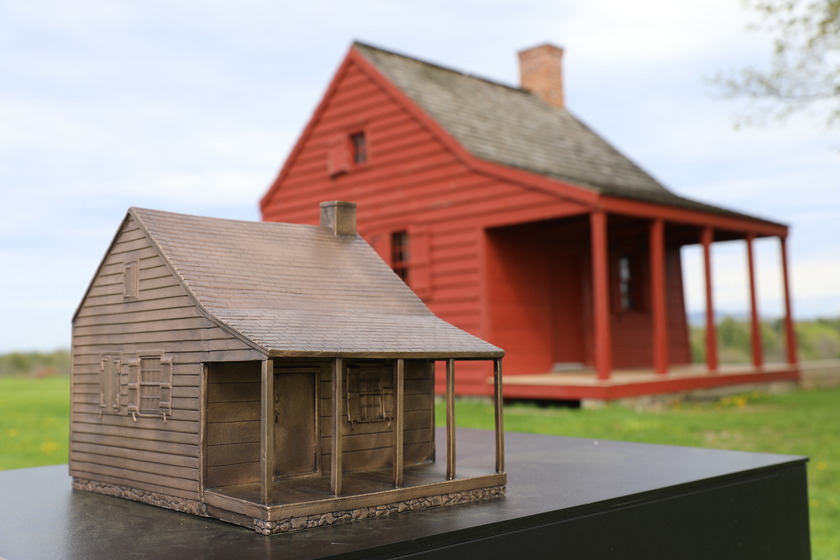 This bronze casting includes a copy of the actual Neilson House structure includes a shingled roof with chimney, horizontal siding, windows with shutters, a porch with four posts, and rocked foundation. The Neilson House, a small red farmhouse, sits in the background on a green, grassy hill.