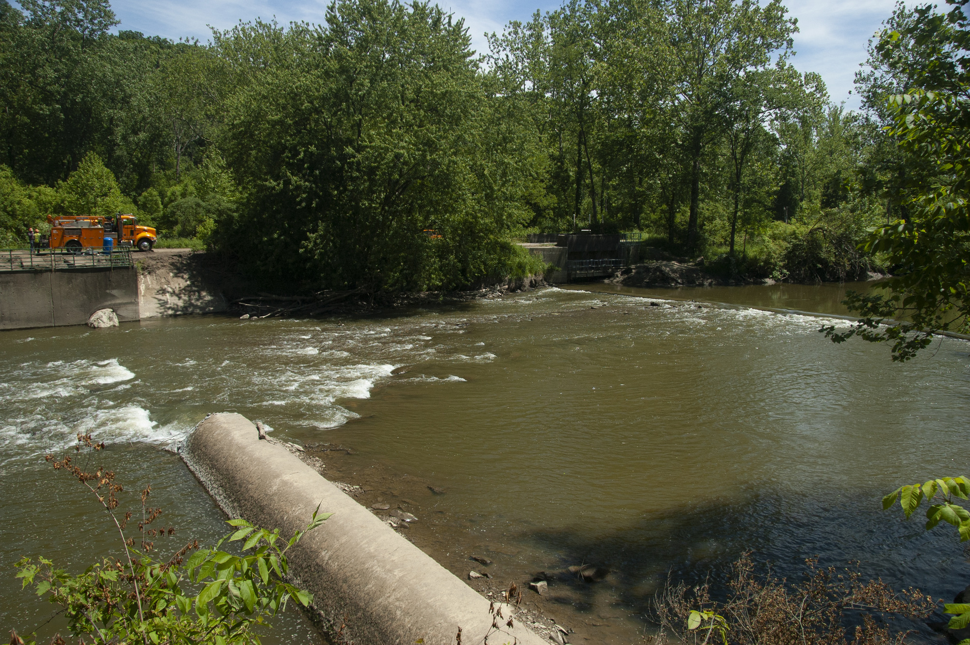 The partially removed Brecksville Diversion Dam is on the left. Also on the left, an orange truck, accompanied by two workers, rests on the road that runs alongside the Cuyahoga River,. The canal-era Pinery Feeder Dam is now visible on the right.