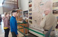 Uniformed map in front of a large wall map with a man and women inside a large room.