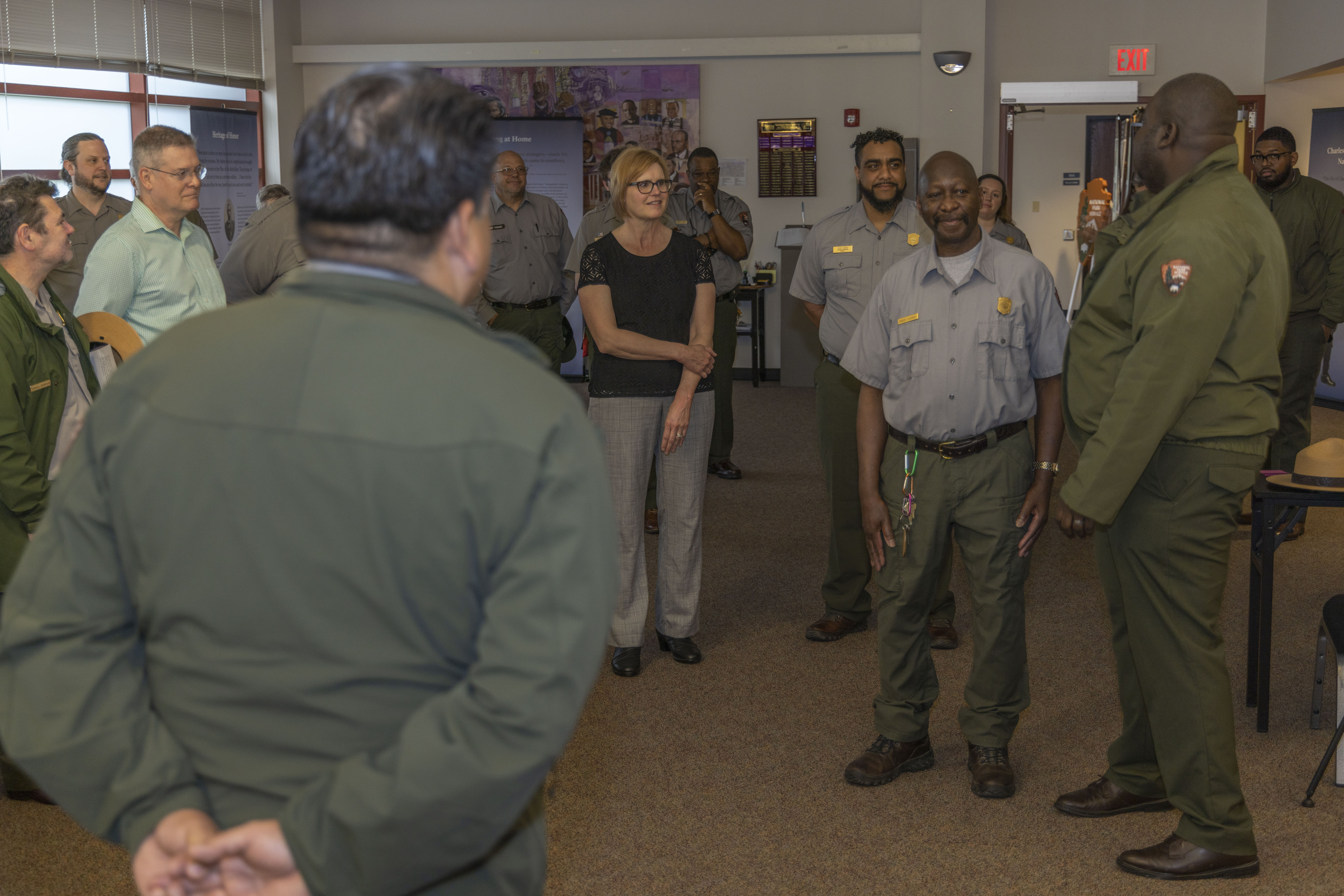 A man in a green uniform with his hands folded behind him listens to a man in a gray shirt speak to him