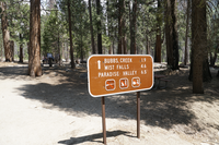 A brown metal sign stands in front of a picnic table surrounded by trees. In the background their is a group of visitors standing by a sign. 