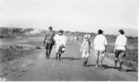 The image is in black and white. A group of 11 or more people are walking, backs facing the camera, down a smooth gravel path. The path takes up a majority of the foreground, tapering off in the distance. Those closest to the camera are in focus, wearing boots, hats, trousers and shirts. From the right side of the image three women are walking side by side. The second woman from the right is identified as Meta Lovejoy. To the left of Meta is a girl. The girl is identified as Charlotte Lovejoy. Charlotte and Meta are holding hands as they walk along the road. The hikers further down the road are blurry. The landscape on either side of the path is sparsely covered with vegetation like grass, shrubs, and trees. Plumes of smoke rise in the distance. In the background, the slope of a shield volcano is faintly visible.