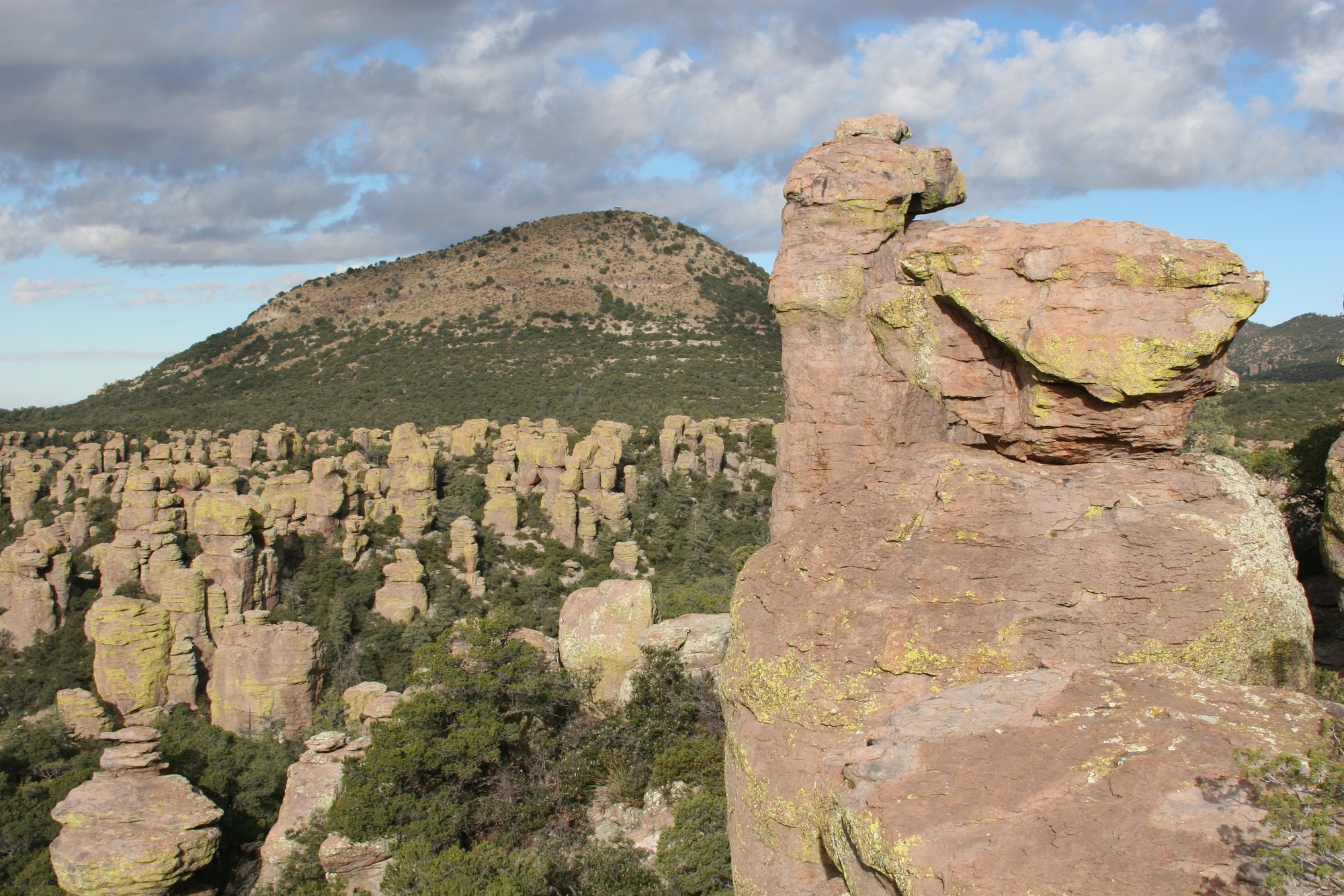 Rock formations in the foreground with a rounded peak in front of a cloud-filled blue sky. 