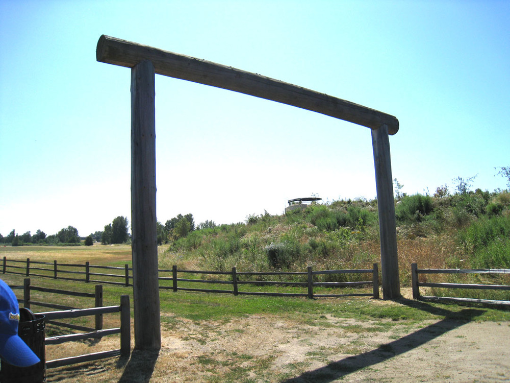 A wooden gate over a dirt road is formed by two vertical logs on either side, spanned by a cross bar on top.