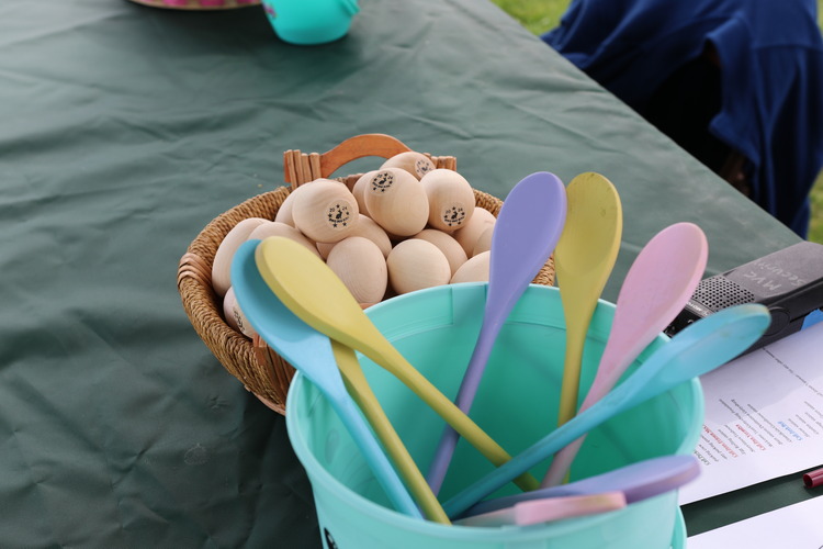 A bucket of painted wooden spoons sits next to a bucket of plain wooden eggs on a table. 
