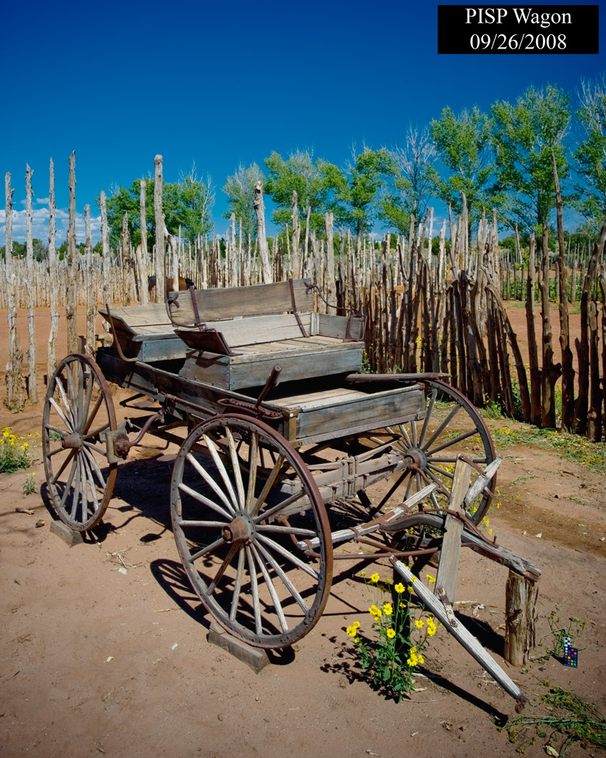 A democrat wagon sits on the grounds on Pipe Spring National Monument. Wooden post fence behind wagon with a horse corral enclosed in the fence. In the background a line of poplar trees can be seen and a dark blue sky overhead.