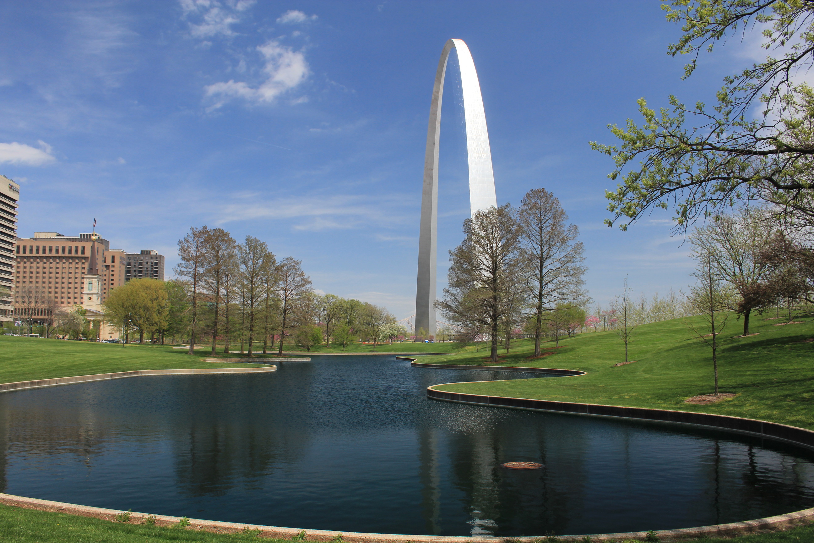 Dark green south pond is in the foreground reflecting the city scape and trees in the water. The pond is surrounded by green grass and the Arch stands in the background surrounded by a bright blue sky and a few white clouds.