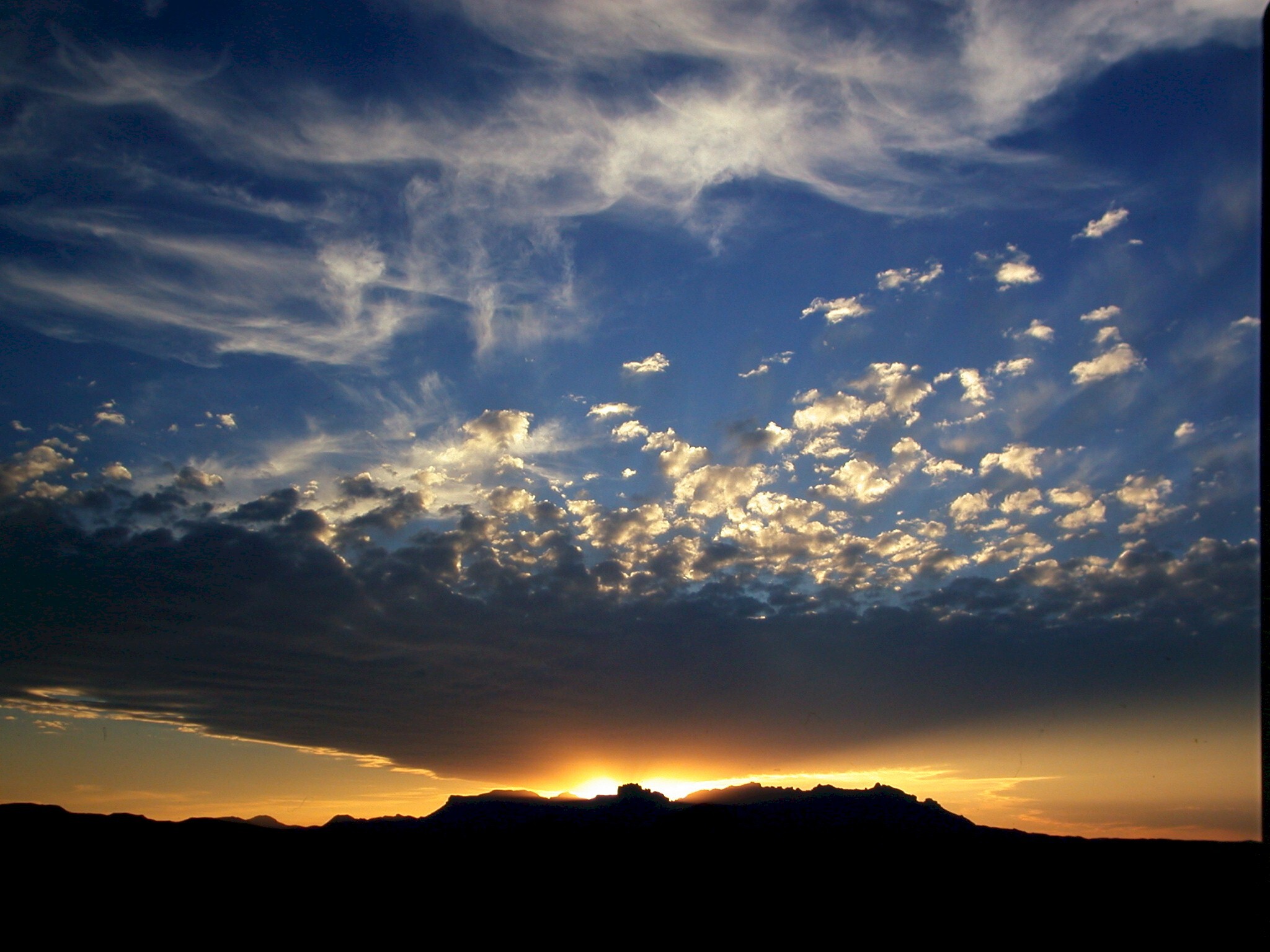 The sun sets behind an isolated mountain range, casting golden light on high, wispy clouds.