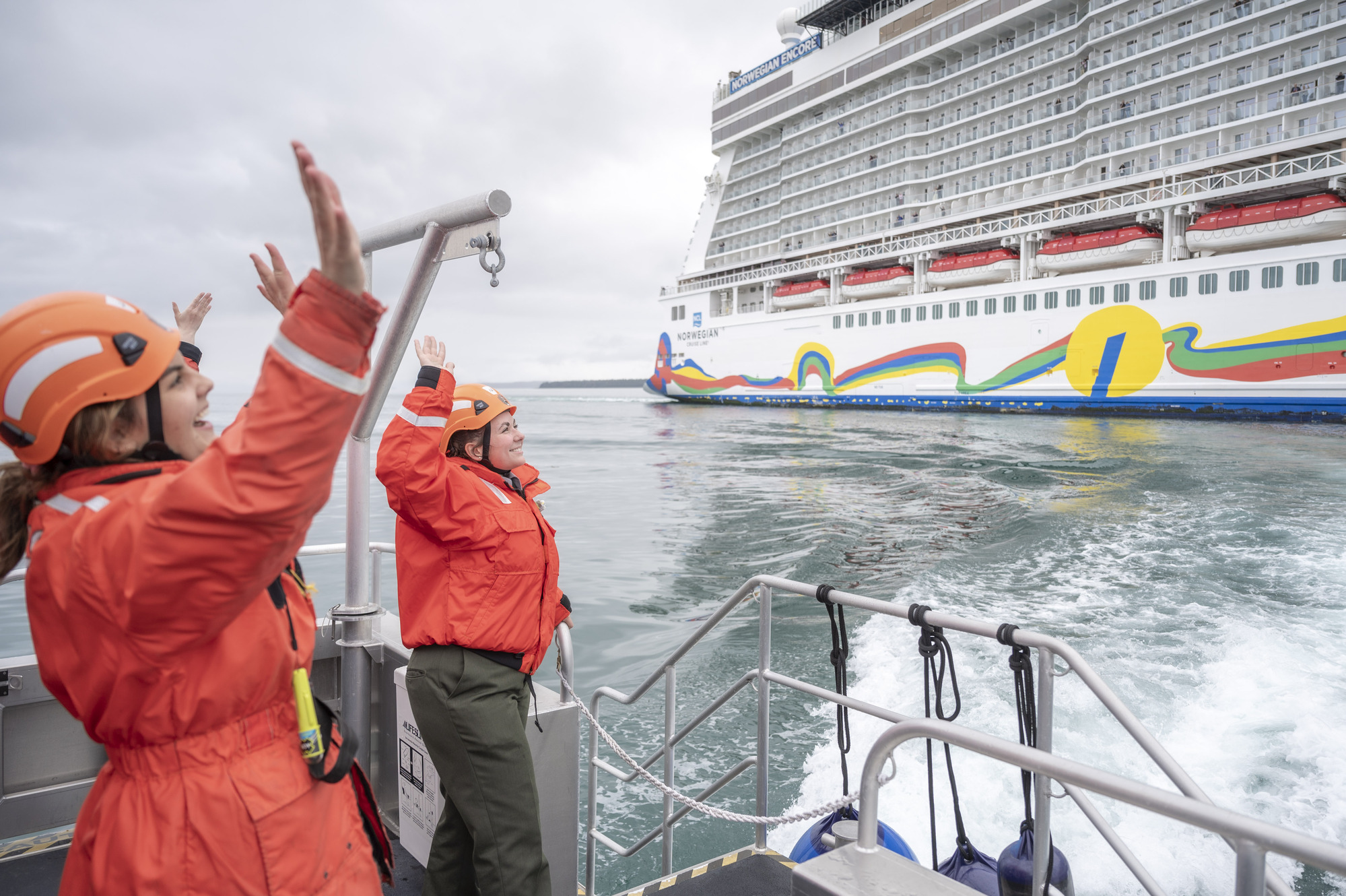 Two park rangers wearing orange safety flotation jackets wave and smile enthusiastically toward a passing cruise ship.