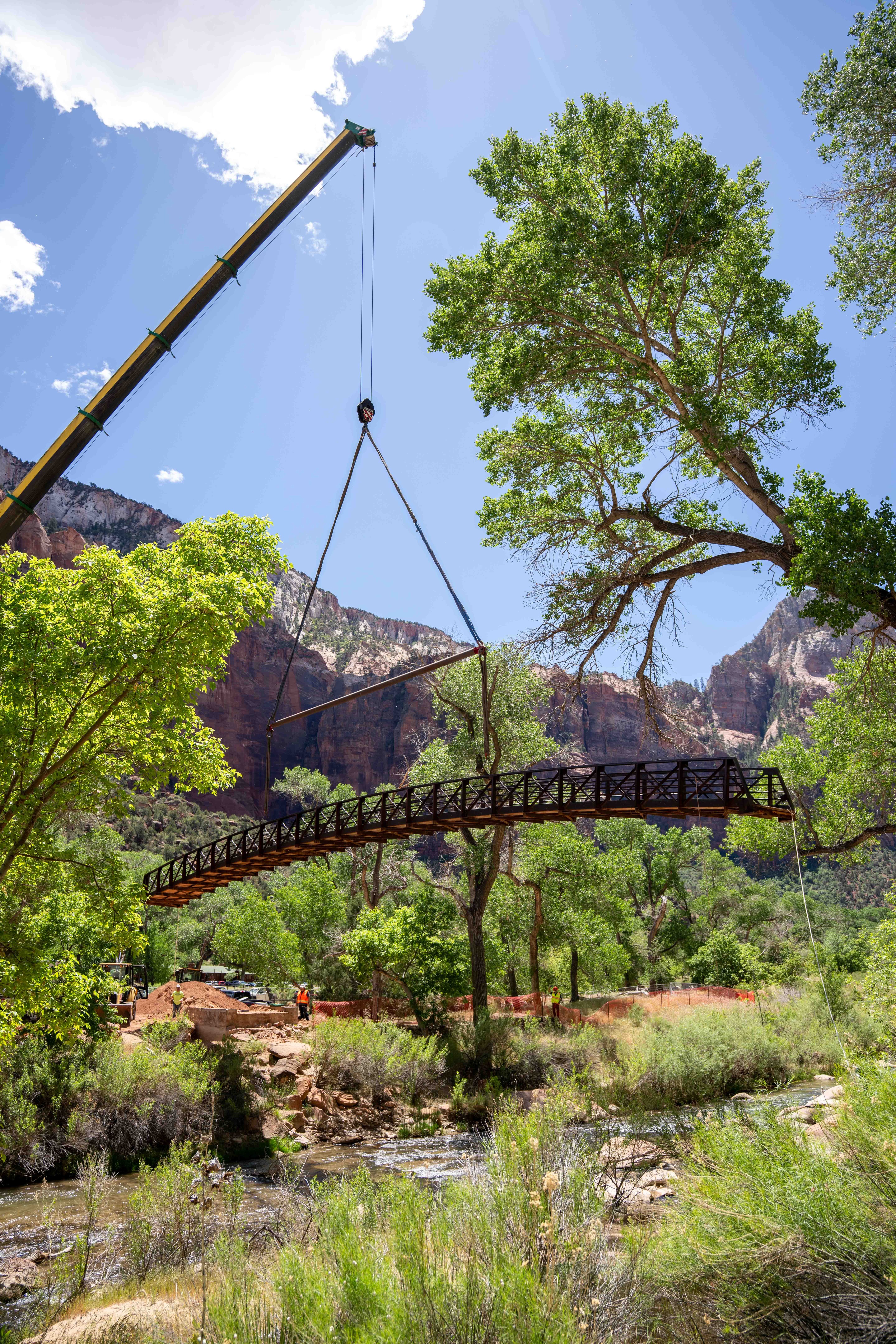 a large crane hoisting a steel bridge in the air with trees and red sandstone cliffs in the background