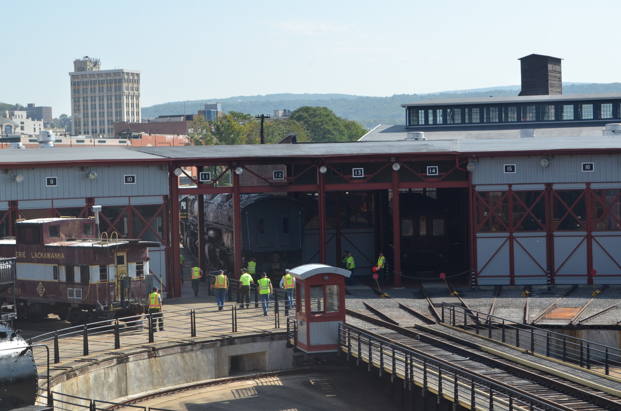 Elevated exterior view of a large train being pulled off a turntable with 10 people looking on. A second train is in a threshold doing the pulling.