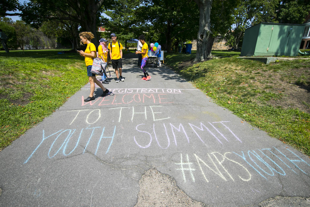 Kids walk along a sidewalk that has "Welcome to the Youth Summit #NPSYouth" written in chalk. 