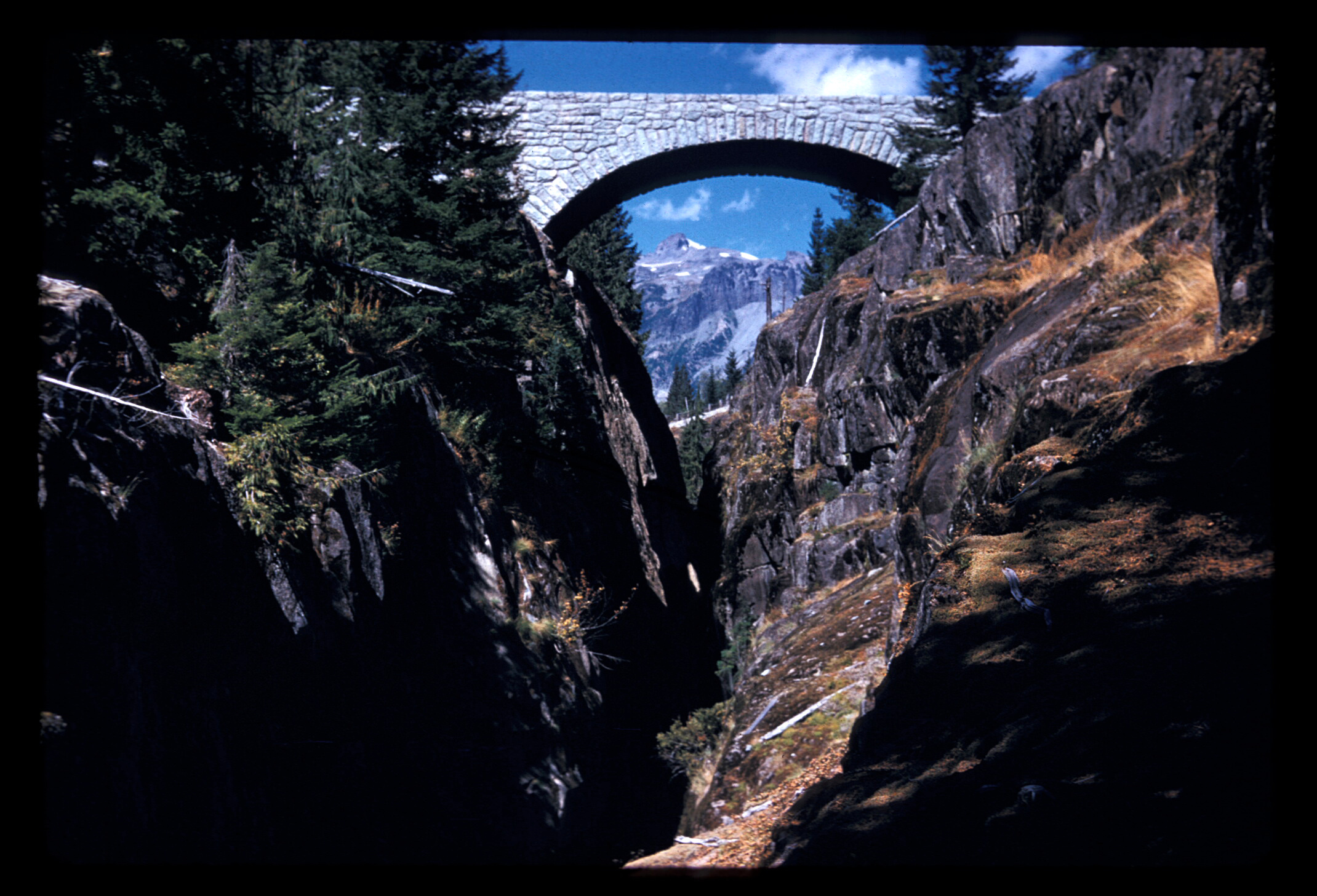 A stone-clad concrete bridge arches over a narrow rocky canyon framing a mountain peak under the bridge's arch. 