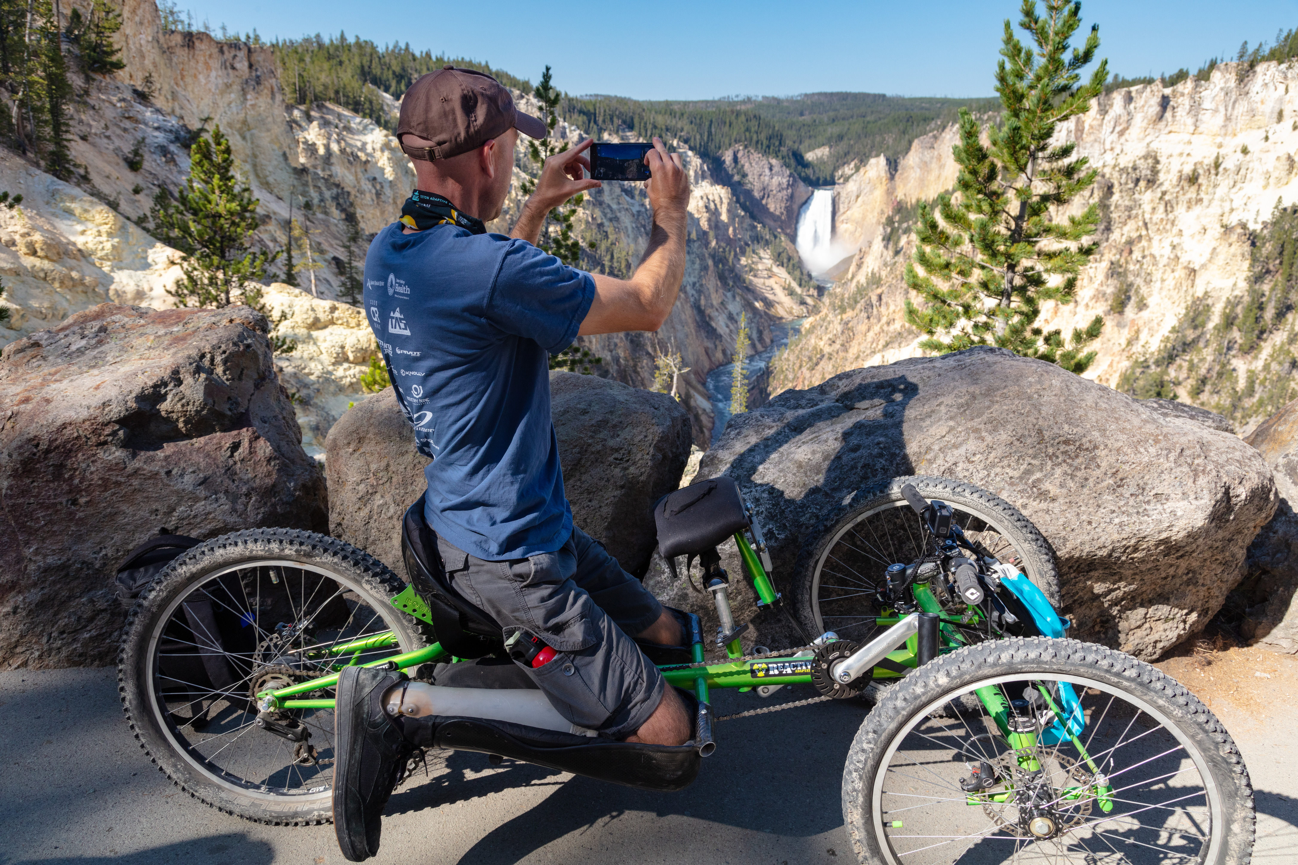 Man sits in three wheeled chair and turns to his left and take a photo of the Lower Falls, which is 308 feet high.