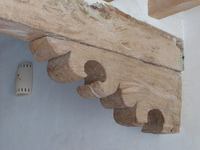 A detailed wooden beam with carved ends near an adobe wall and a small ventilation duct.