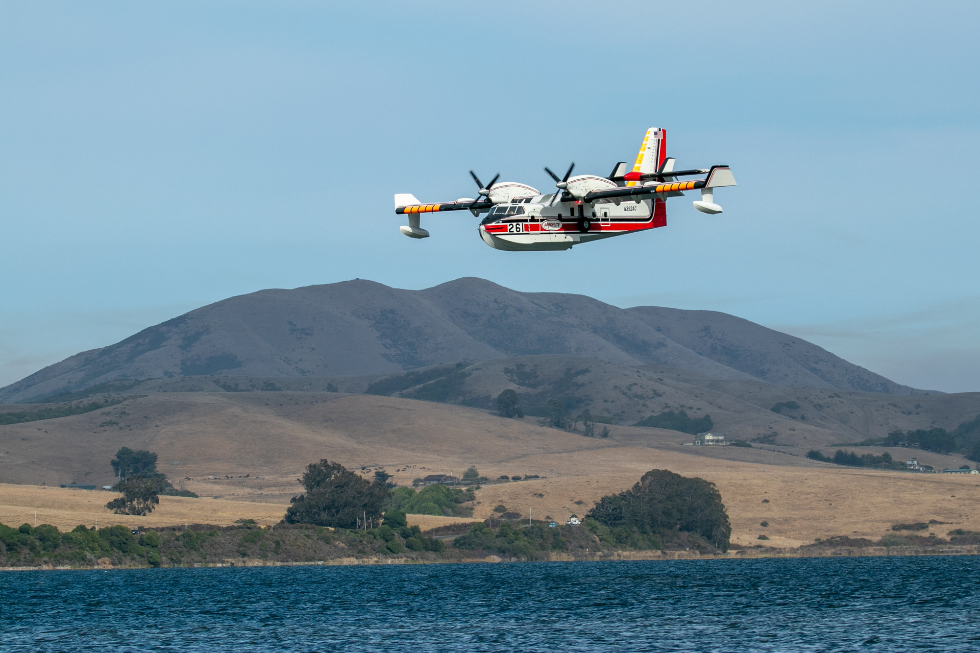 A twin-propped white plane with red and yellow markings flies low over a narrow bay with tan-colored hills in the background.