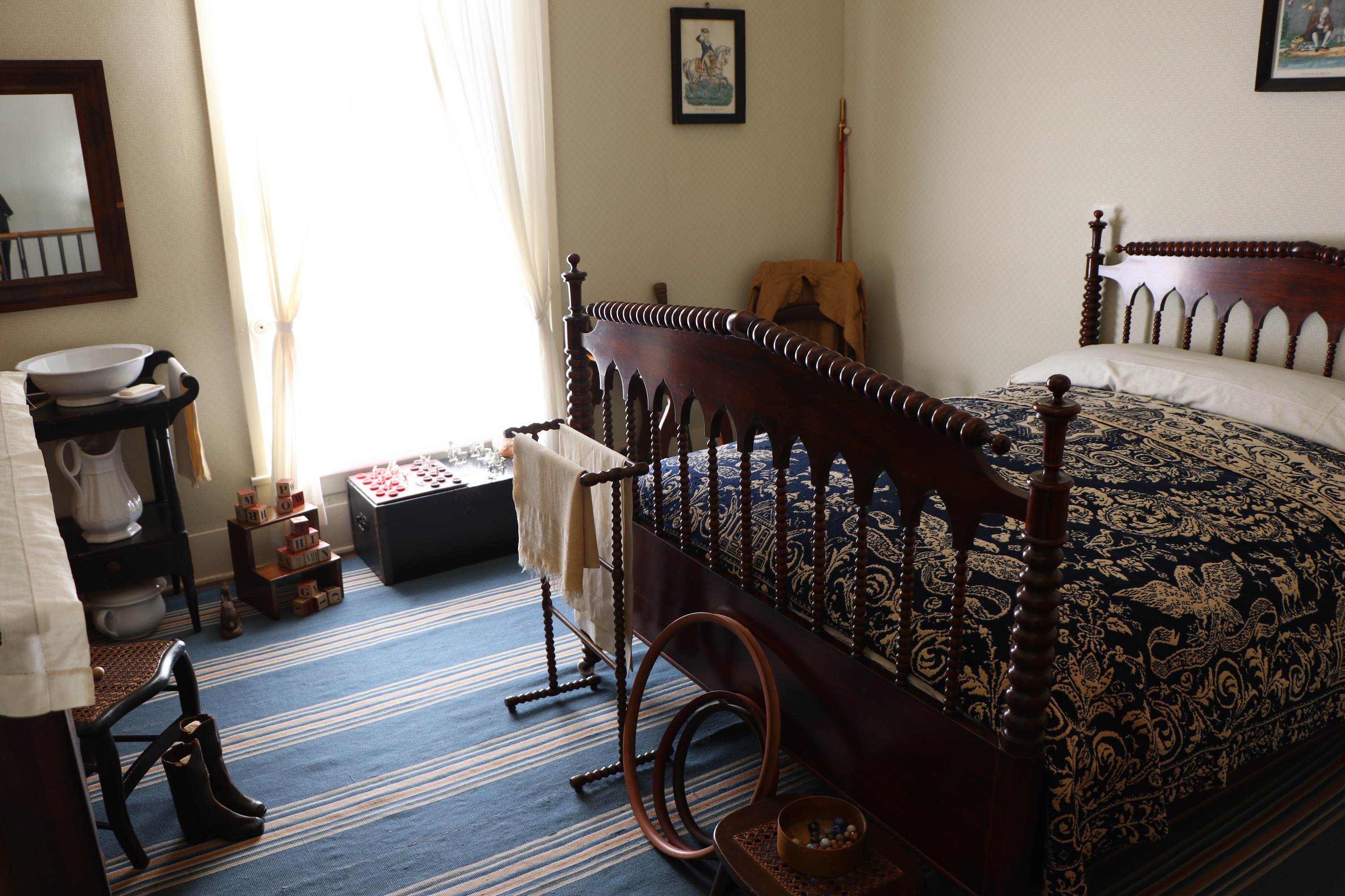 A bedroom with a blue striped rug, plain walls, and historic toys from the 19th century.