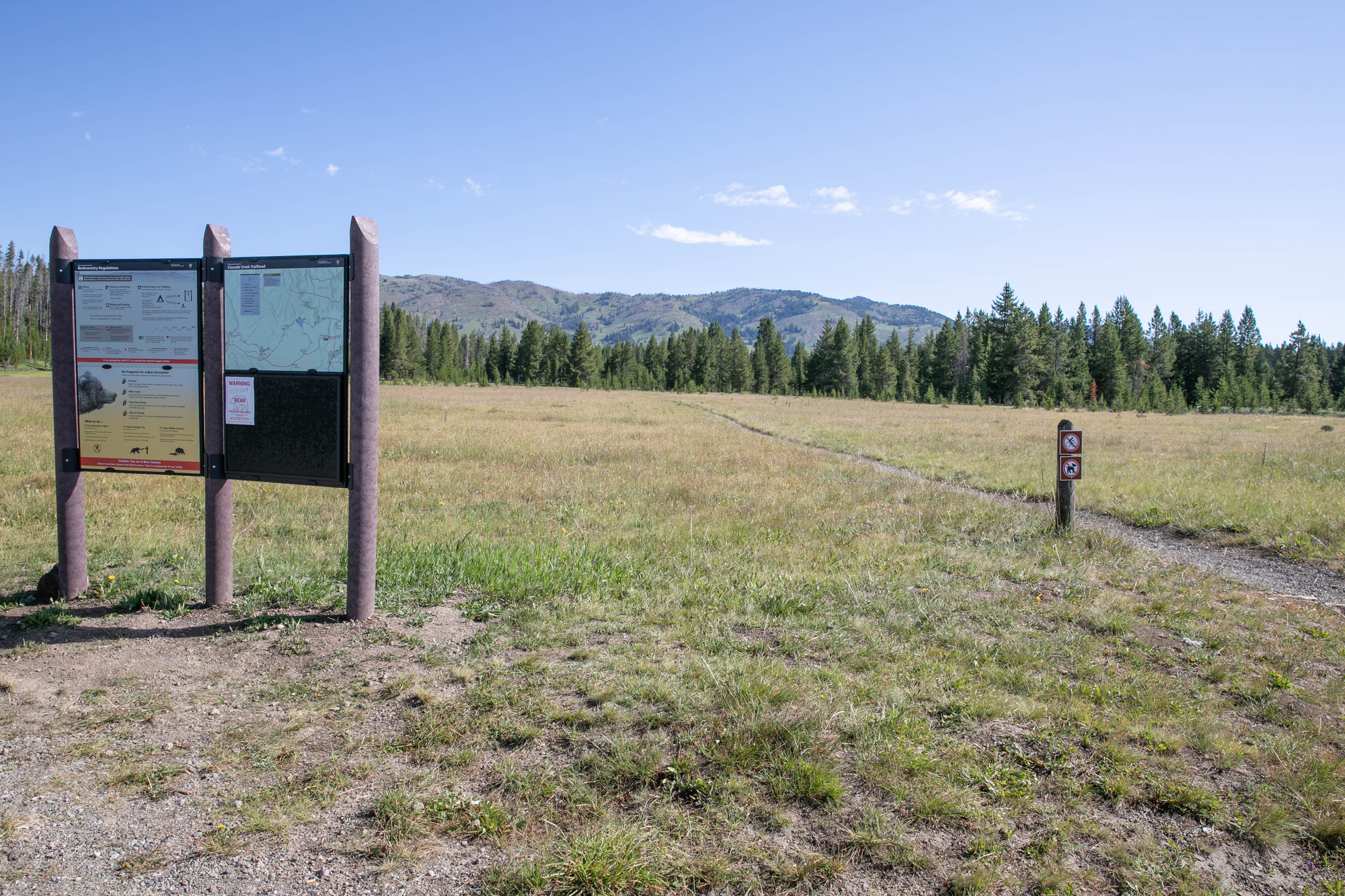 Trail passes a large trail sign board to the right and crosses an open, grassy meadow.
