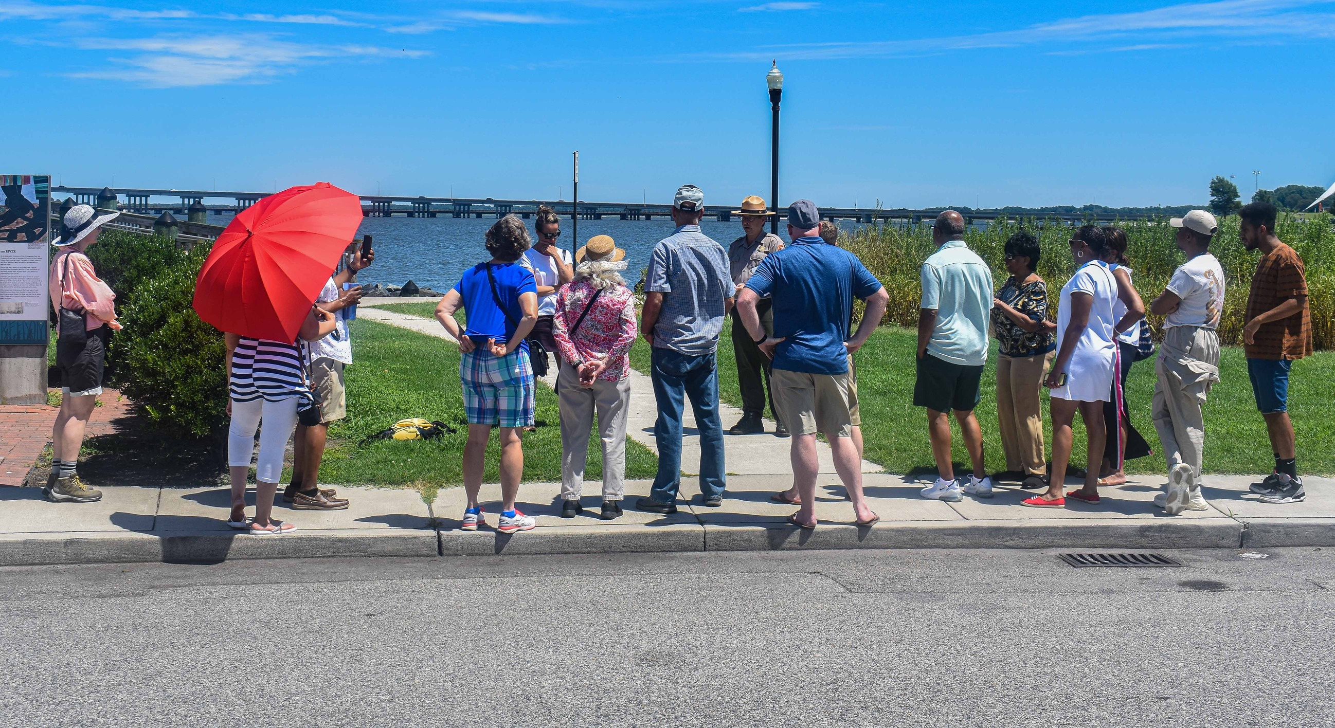 In the foreground, an asphalt road can be seen. An NPS Ranger can be seen presenting a piece of paper while giving an interpretive talk to a group of 13 guests standing on a sidewalk at historic Long Wharf in Cambridge, MD.  A light post, the Choptank River, and the Choptank Bridge can be seen in the background.