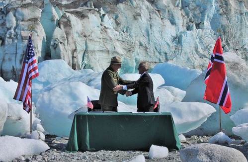 Two men shake hands in front of a field of small iceberg boulders with the flags of the US and Norway on either side of them.
