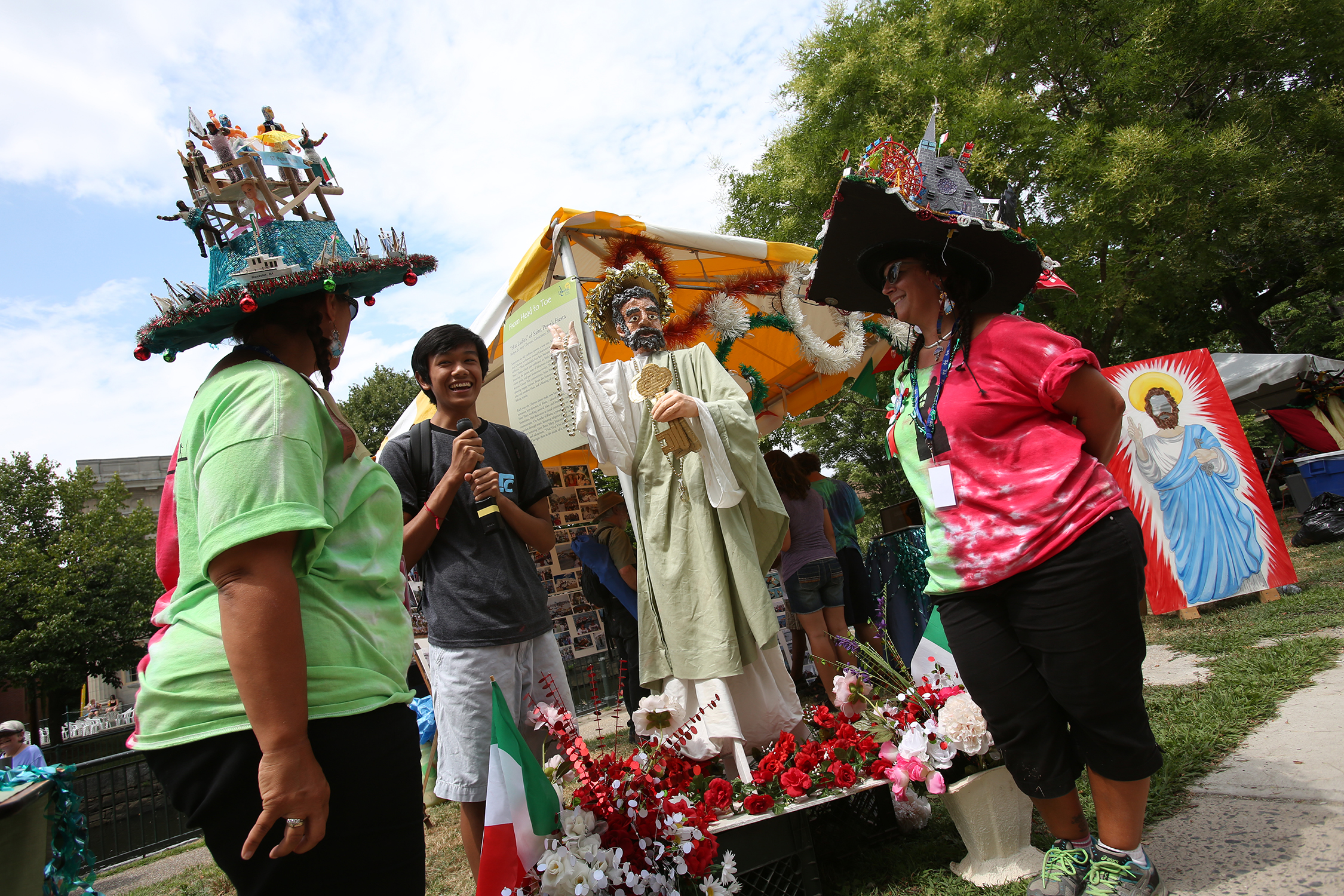 Two women are being interviewed by a young reporter. Their tent has wreaths hanging from it, flower planters displayed in front of the St. Peter statue. Visitors crowd their displays, as a face-cutout of St. Peter is propped up. One woman's hat has a ferris wheel near a mountain (which is the crown of the hat). While the other hat has a band atop a dock, as the crown of the hat is water, and a yacht is on the brim. 