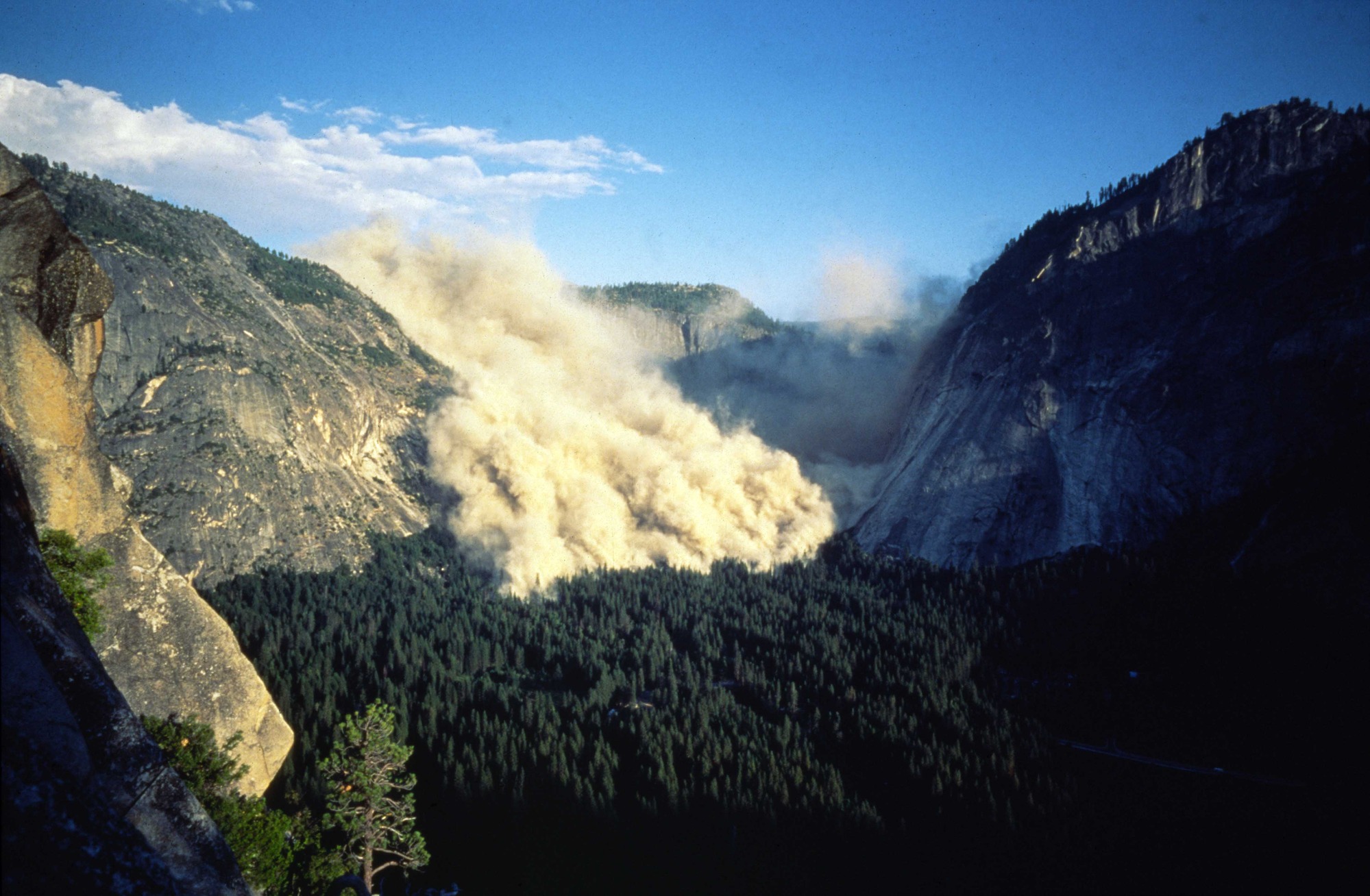A gray dust cloud rises above the tree covered valley floor and fills the end of the valley. Steep rock cliffs rise above the valley floor on both sides.