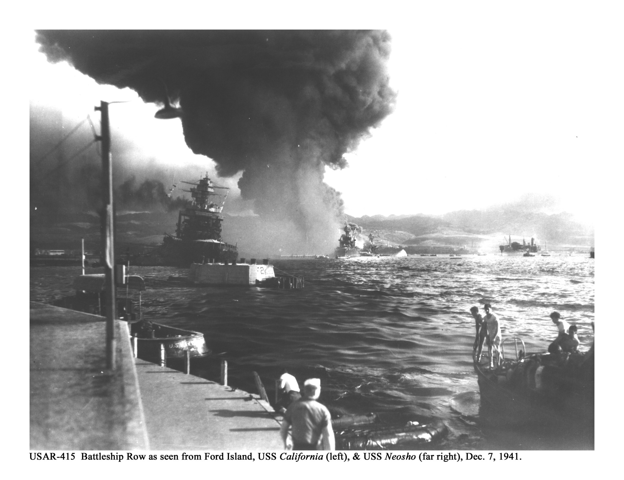 Historic photo of attack on Pearl Harbor, looking out into harbor from land. Sailors aboard lifeboats in foreground and battleships engulfed in flames in background.