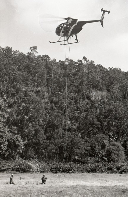 A black and white image of a helicopter in the sky with a rescue rope dropped. Towards the bottom left corner are two people walking towards the rope that is hanging from the helicopter in the center of the image.