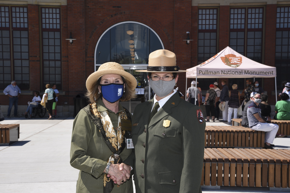 Two women shake hands in front of the Administration Clock Tower Building. The woman on the left has blonde hair and wears a straw sun hat, dark blue mask with IDNR logo, and green jacket with a gold patterned scarf. 

The woman on the right wears a formal National Park Service uniform dark green jacket and straw hat with a grey face mask. Behind them, people walk past and sit on benches. 