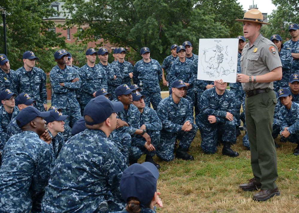 a large group of men and women in Navy uniforms listed to a male park ranger. 