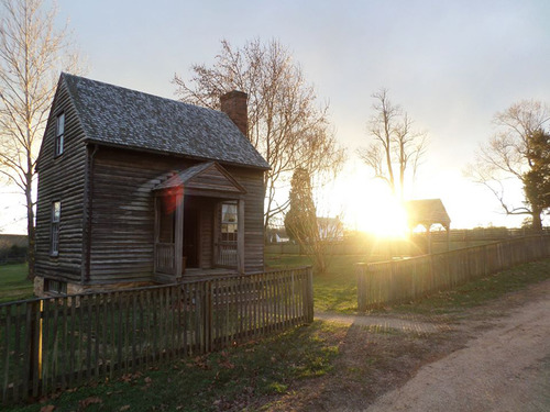 A small wooden two story house with a fence between the house and the road with the sun on the horizon to the right of the house.