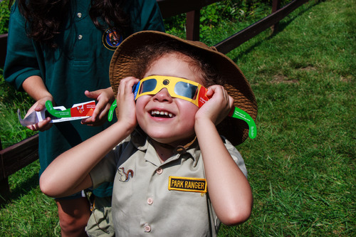 A smiling young girl in a junior ranger uniform watches the eclipse using special glasses. 