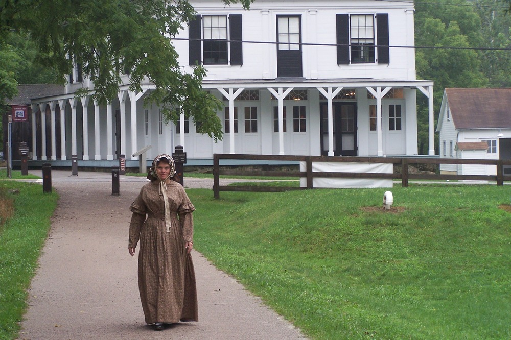 A woman in modern times wearing an historic long skirt and bonnet walks along a dirt path away from a two-story white building with wraparound porch.