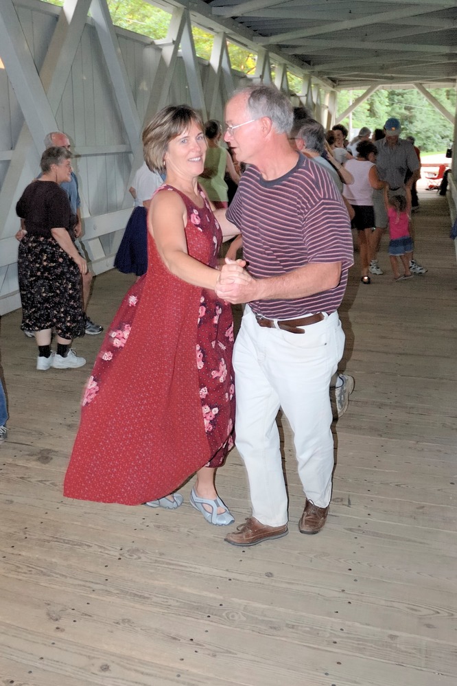 A man and woman dance with other dancers in a wooden covered bridge behind them.