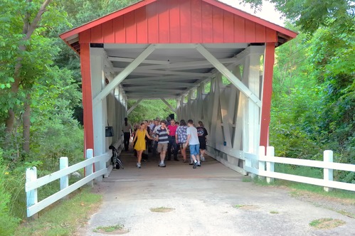 Several people dance holding hands down the center of a red wooden covered bridge with white interior.