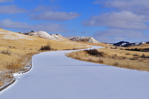 Snow covered road running through dry prairie grasses.