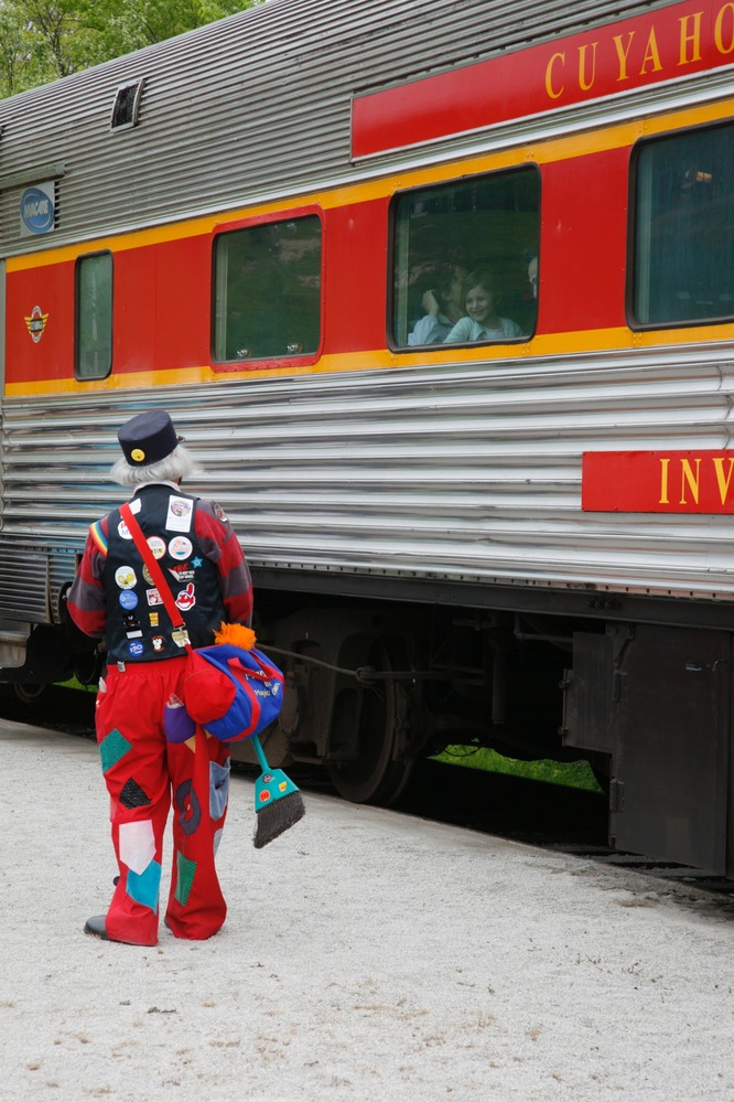 A person in red pants and black vest with many colorful patches faces a silver and red train car; children in a window smile down at them.