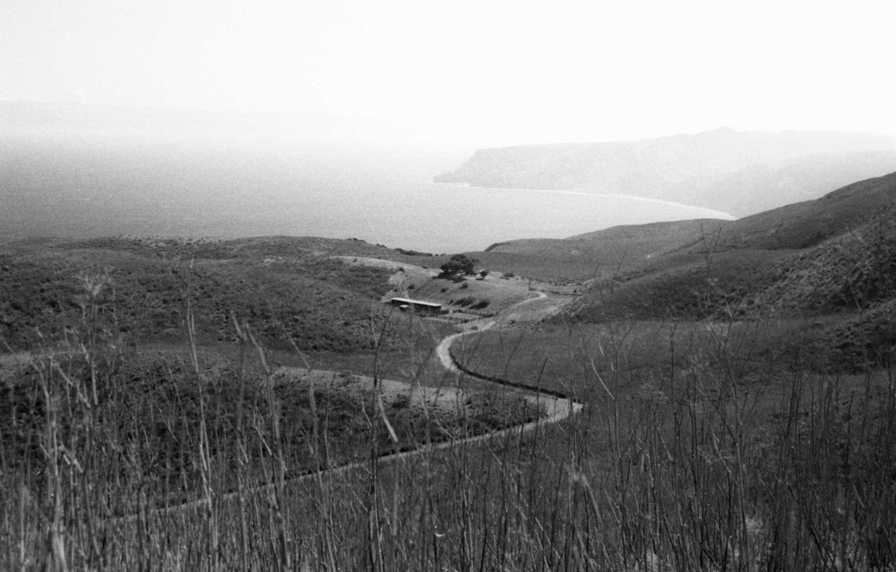 A road winds through rolling, open terrain near the coastline. The ranch is protected by the folds of the land.