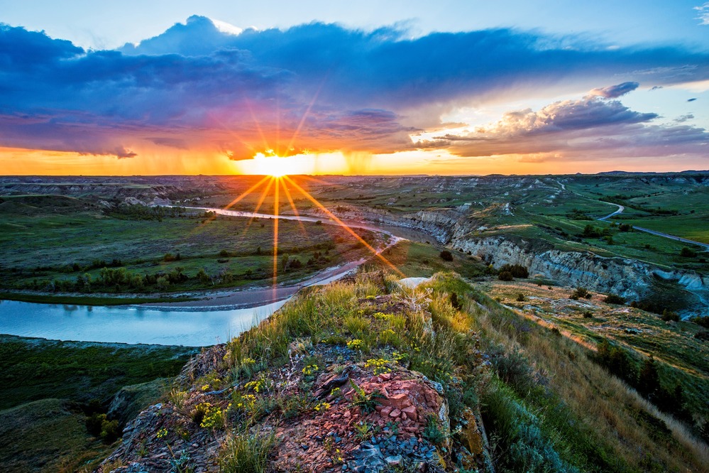 Rays from a setting sun burst through cloud to shine on a rocky butte and river down below.