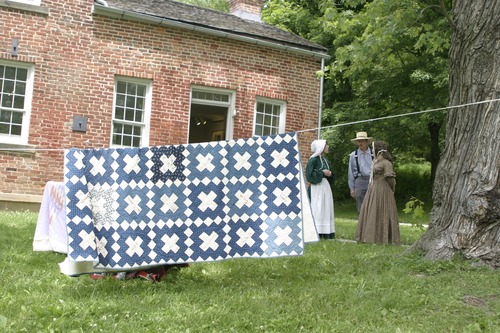 Three people in period dress stand outside a brick building next to a blue and white quilt hanging on a laundry line.