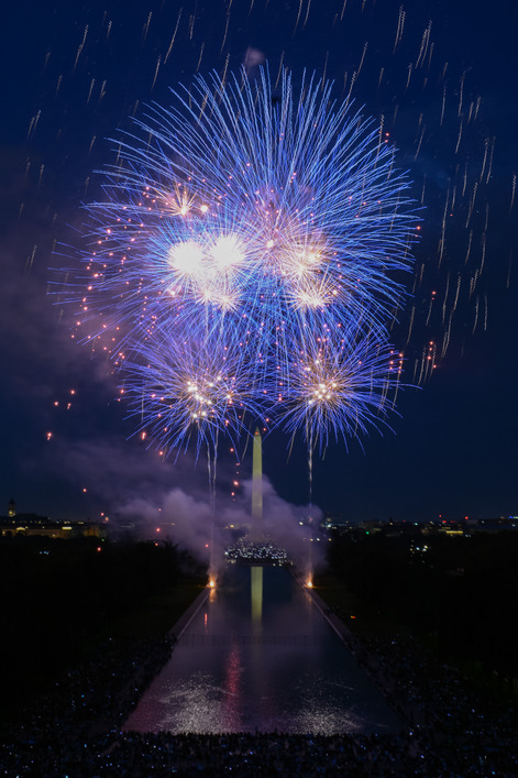 A reflecting pool at night leading towards the Washington Monument where blue fireworks are exploding in the sky.