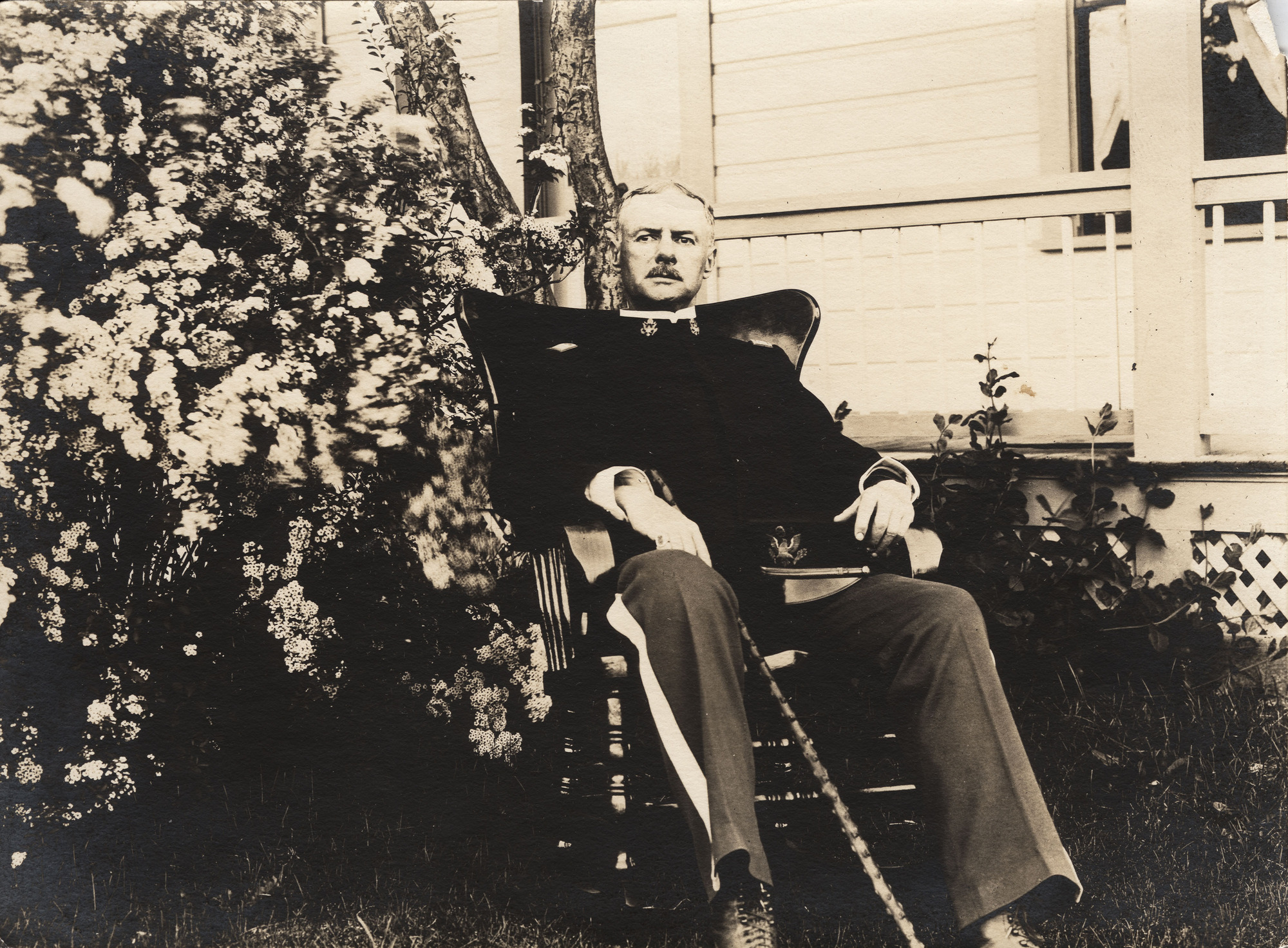 Black and white photograph of a man in a uniform sitting in a chair in yard by a house