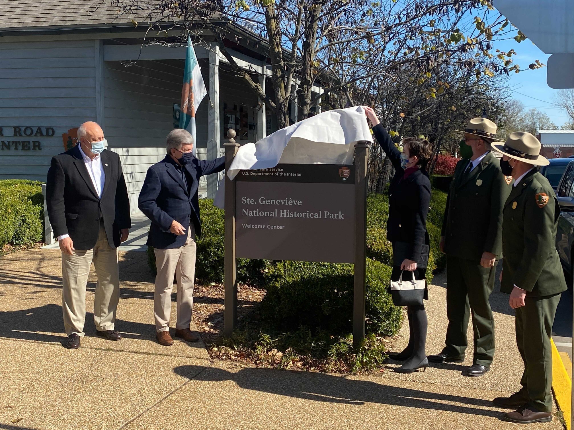 A gentleman and lady dressed in winter attire and face masks remove a white cloth from a sign, as two park rangers and another suited man watch.