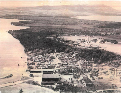 A small town on the edge of a large bay. In the background, separated from the town by a buffer of trees, is two large buildings in the middle of a field.