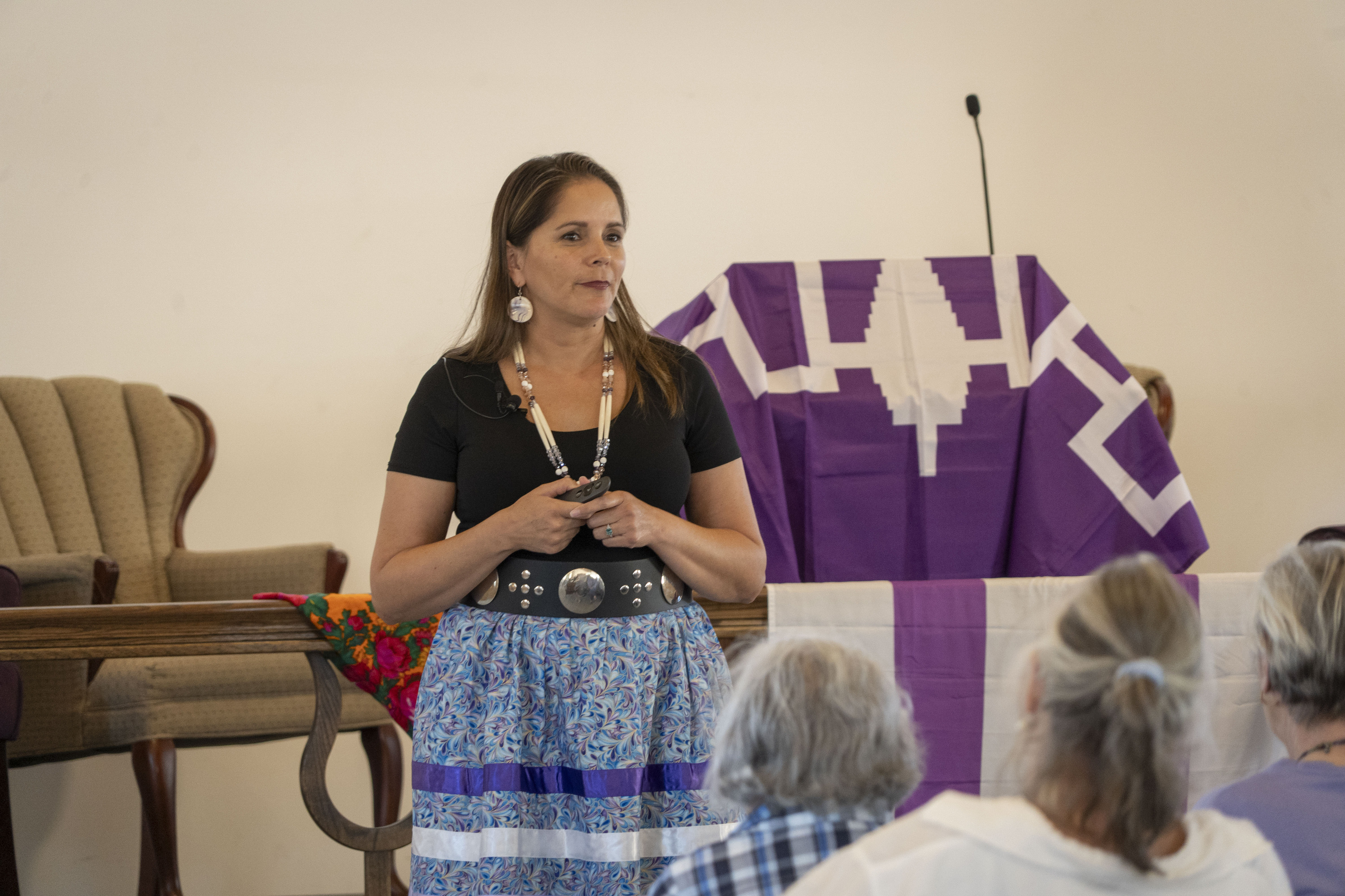 A woman in a blue and purple ribbon skirt stands in front of a Haudenosaunee flag