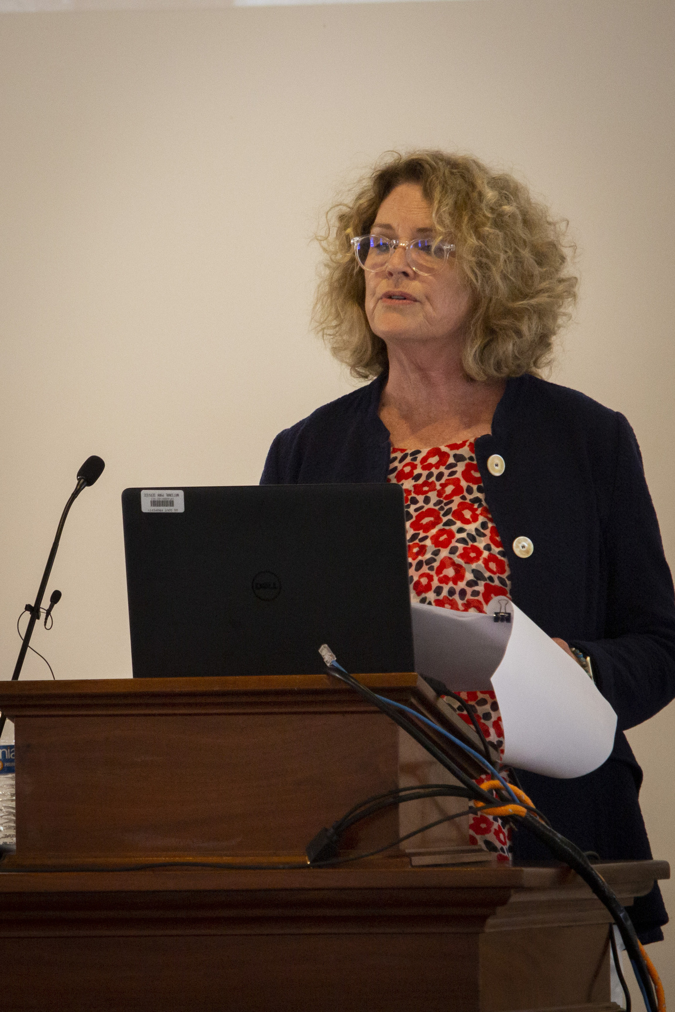 A woman wearing glasses stands and reads from a computer.