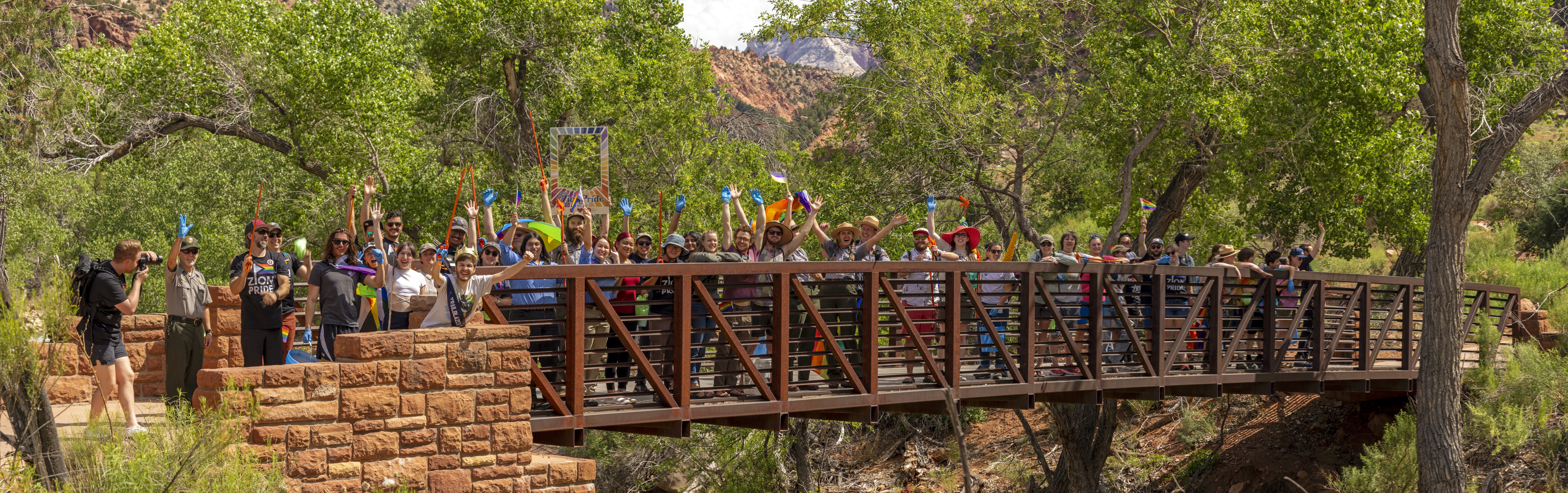 A large group of people in colorful clothes and gear stand on a bridge over a river, smiling and waving at the camera.