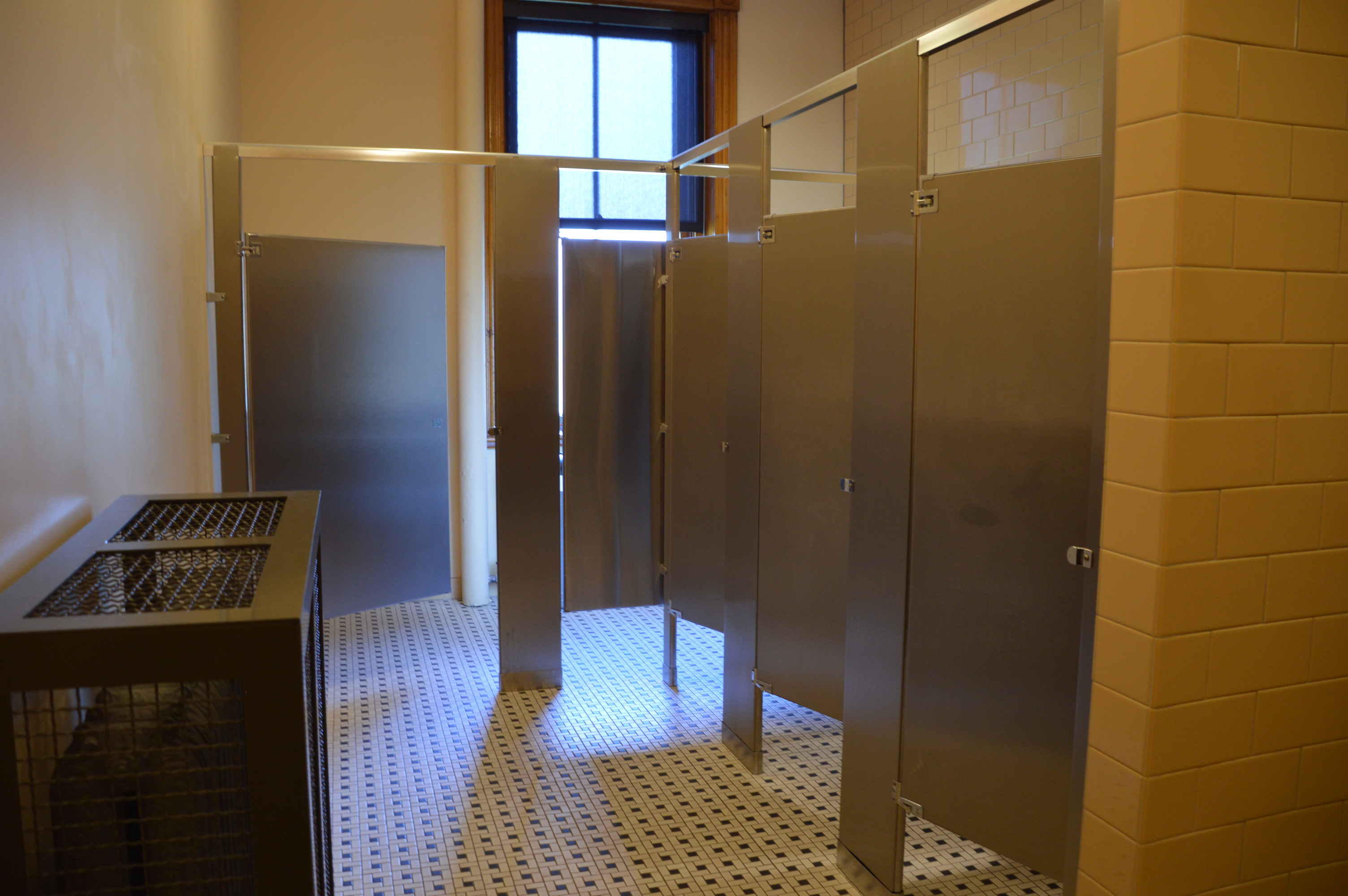 A row of three stalls with metal doors and dividers are on the right. The accessible stall is directly ahead. A caged radiator is in the foreground on the left. The width between the radiator and the dividing wall is 46.5 inches. 