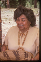 A woman in a Native American attire holds a woven basket.