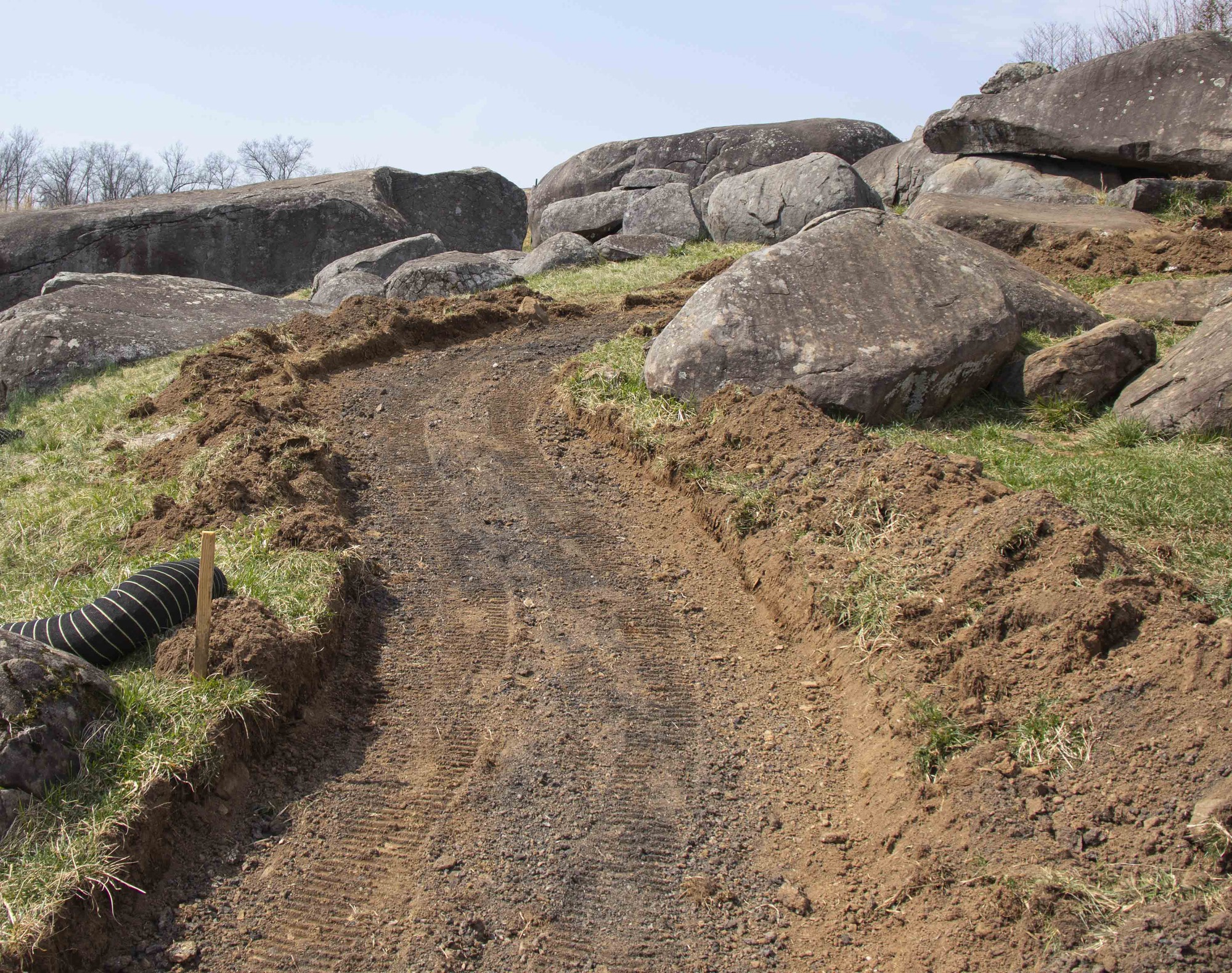 The focus of the frame is a dirt path with its pavement recently removed curling up and to the right of the frame. The path is indented several inches into the soil and has visible tire tracks along its length and piles of soil along its sides. On the right side are a few large rocks. In the background are very large boulder formations.